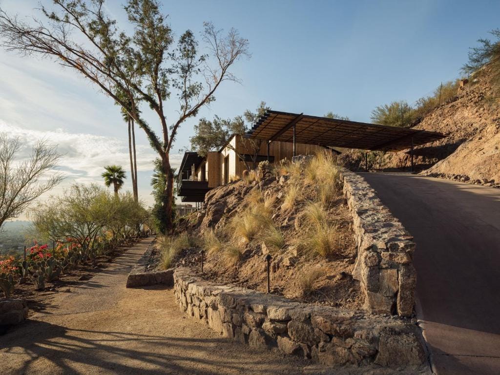 A hillside home with modern architecture and a wooden carport, nestled in a desert landscape with stone retaining walls, native grasses, cacti, and tall trees under a clear blue sky in Teitipac, Mexico.