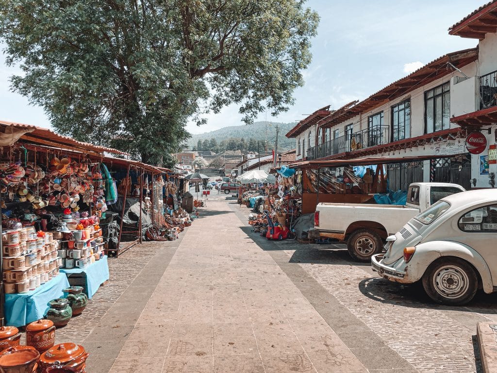 Open-air street market in Tzintzuntzan, Mexico, with artisanal pottery and crafts on display under red-tiled roof stalls, and classic cars parked along the pedestrian pathway.