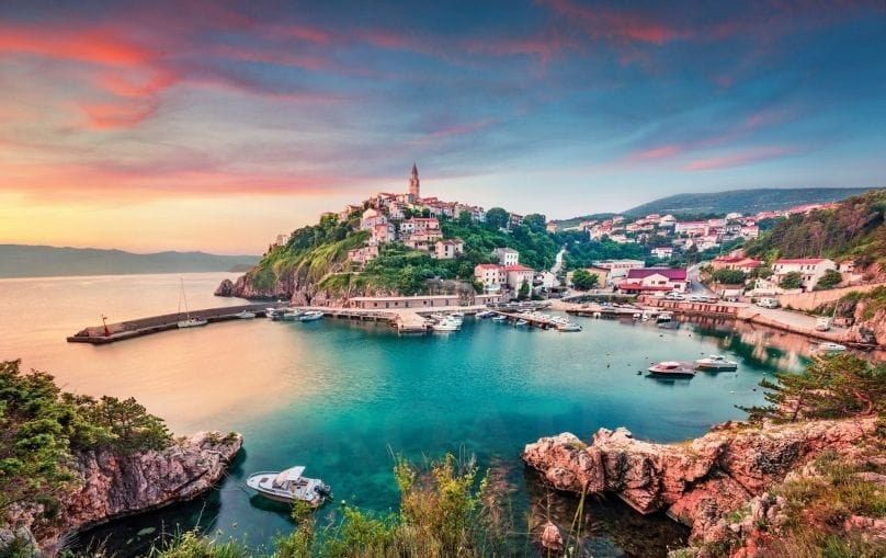 Sunset view of Vrbnik, Croatia, featuring colorful houses clinging to a steep hill overlooking a peaceful harbor with small boats, surrounded by rocky cliffs and a glowing sky.