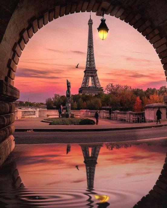 Eiffel Tower at sunset framed by a stone arch, with its reflection rippling in a puddle in Paris, France.