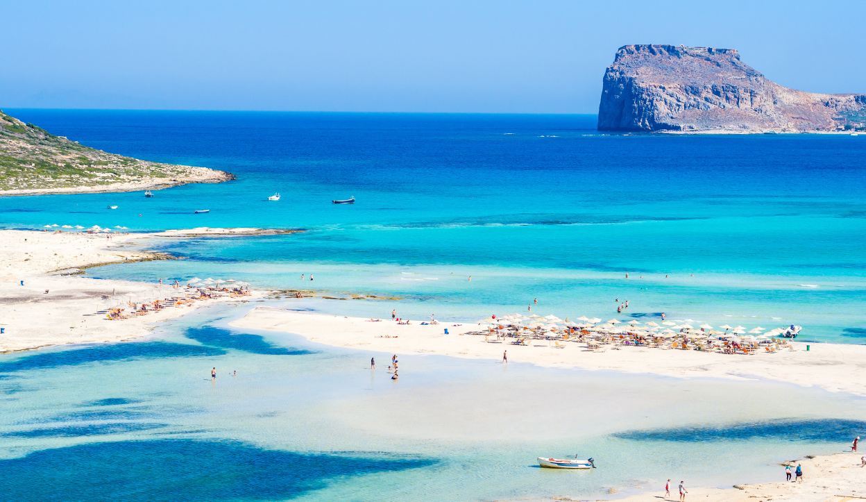 Tourists enjoying the shallow turquoise waters and white sandbanks of Balos Beach in Crete, Greece, with a rocky island formation in the background.