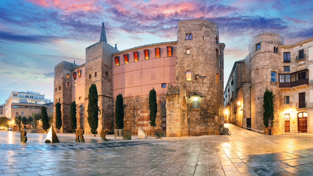 Historic buildings and ancient Roman towers in Barcelona’s Gothic Quarter at sunset, with "BARCINO" sculptures in the foreground.