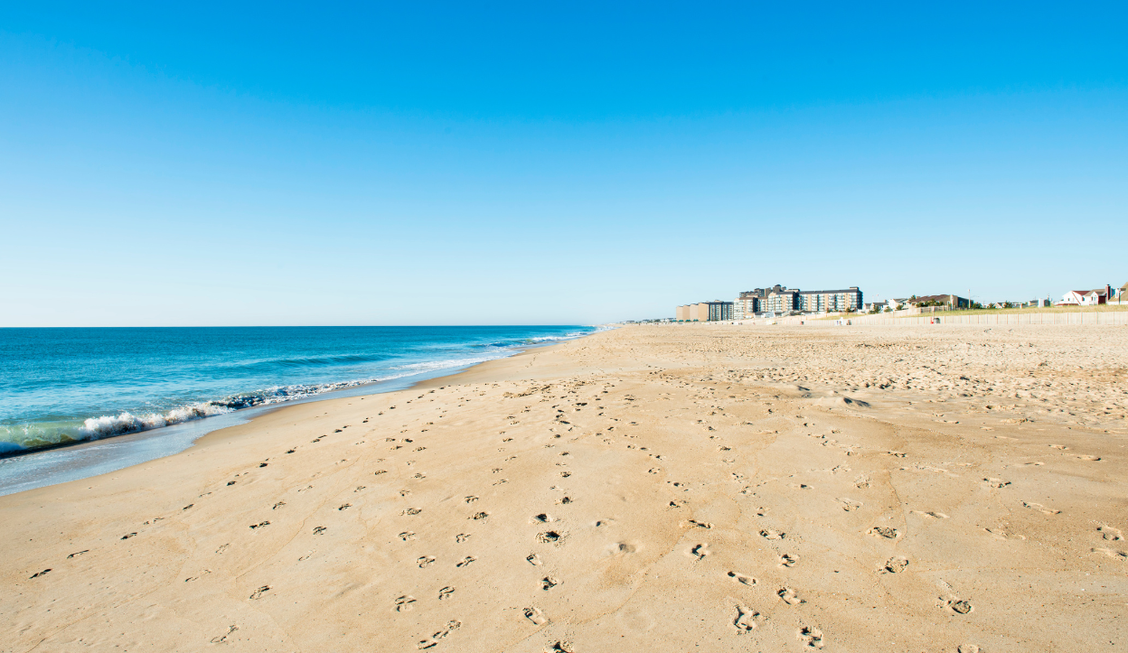 Wooden boardwalk ramp leading to Bethany Beach, Delaware, with beachgoers relaxing near the water on a sunny day.