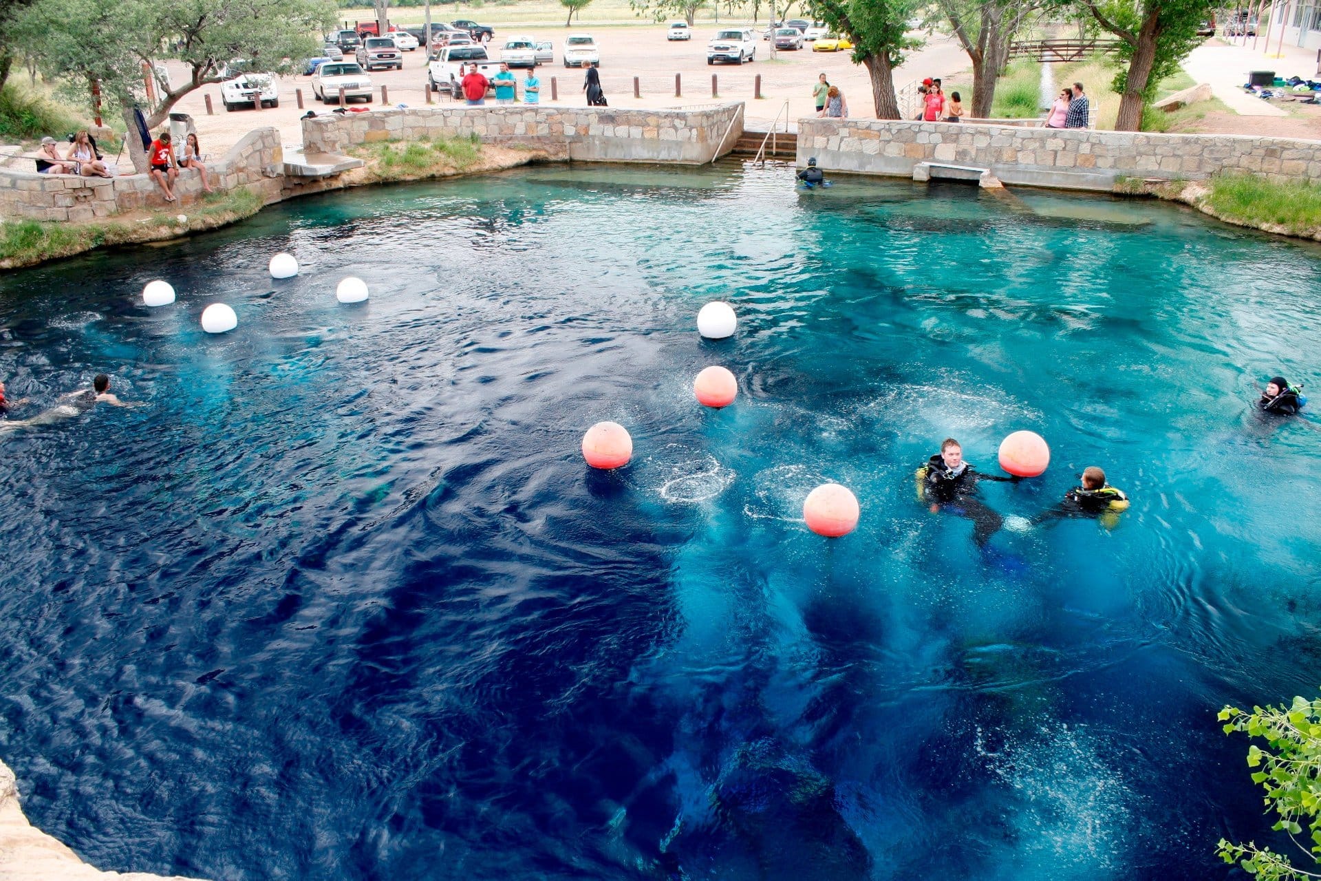Deep circular spring pool known as the Blue Hole in New Mexico, with divers in the vibrant blue water and visitors gathered around the stone-edged perimeter.