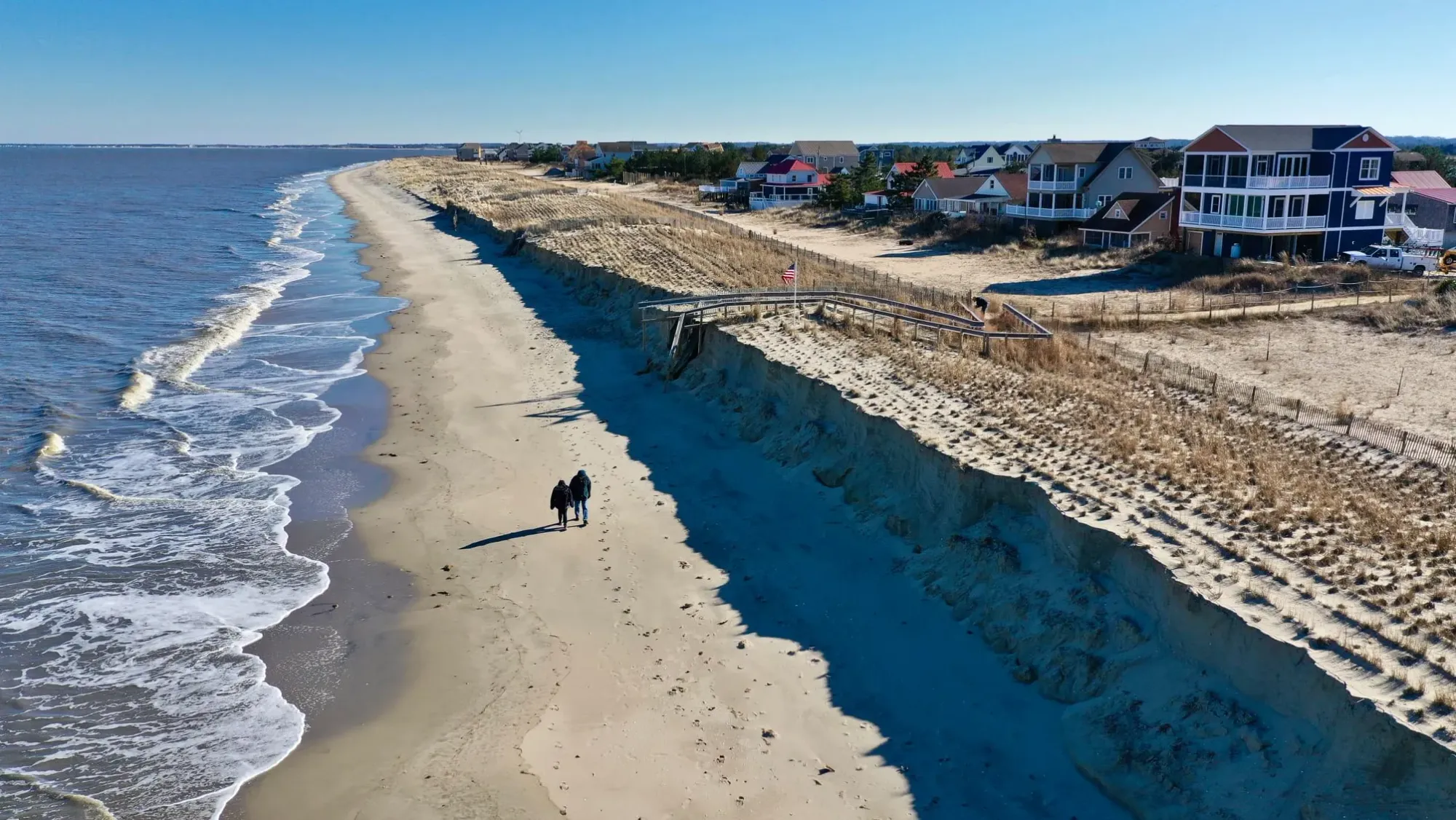 Elevated view of Broadkill Beach, Delaware, showing a sandy shoreline, calm waves, and two people walking along the beach near coastal homes.