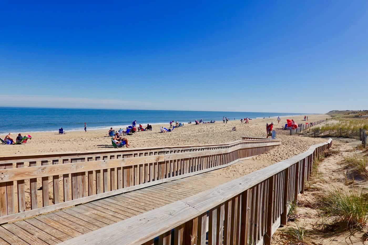 Sandy trail with dune fencing leading to Cape Henlopen Beach, Delaware, overlooking calm turquoise waters and a lighthouse in the distance.