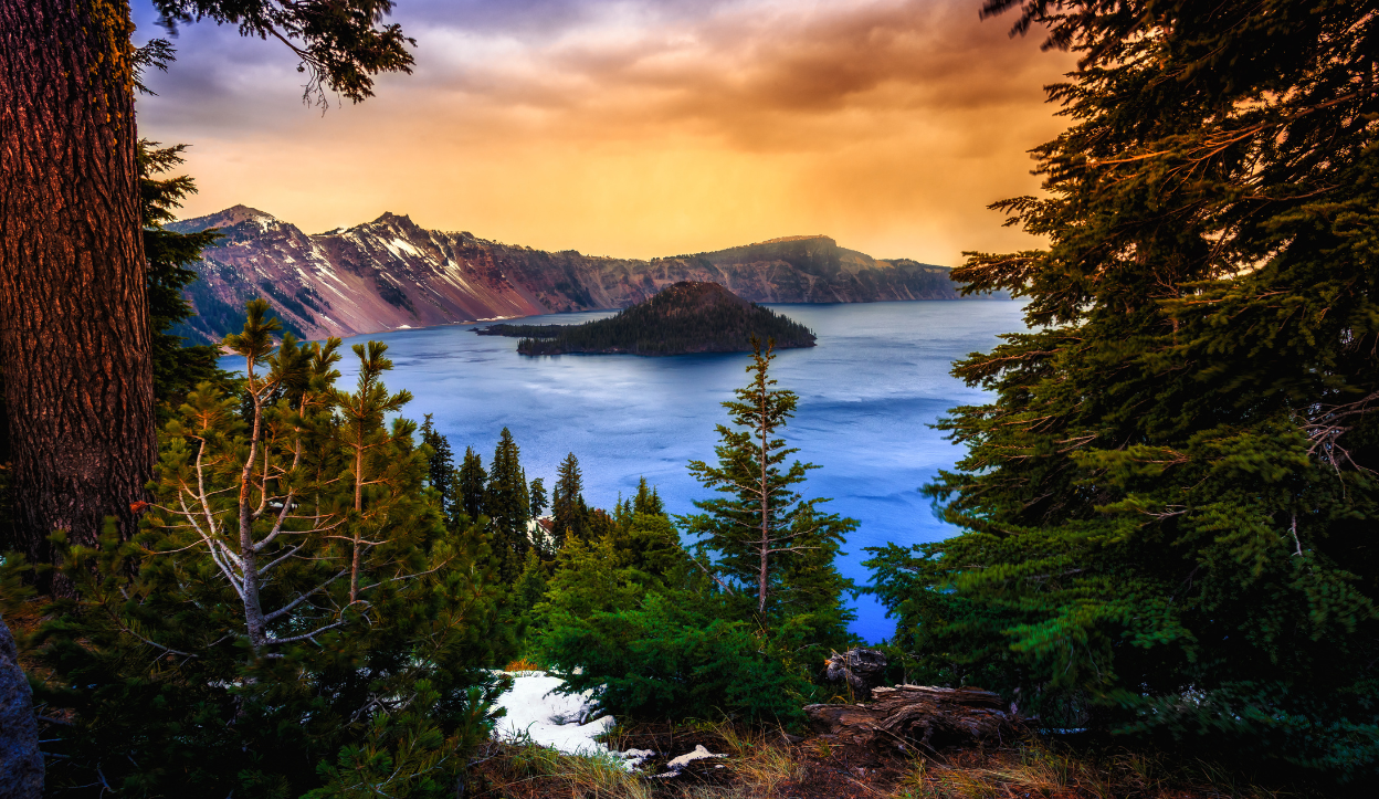 Scenic view of Crater Lake National Park in Oregon showing the deep blue lake, Wizard Island, and snow-dusted mountain ridges framed by evergreen trees at sunset.