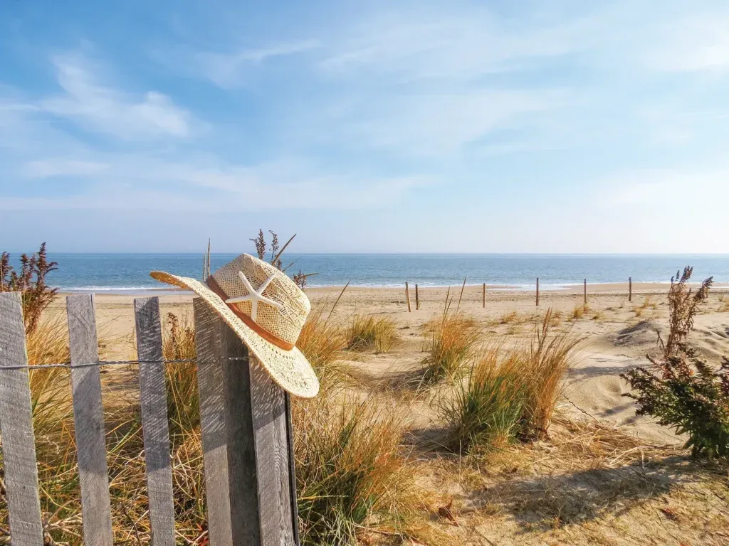 Sandy beach at Dewey Beach, Delaware, with a straw hat hanging on a fence, grassy dunes, and a calm ocean horizon under a blue sky.