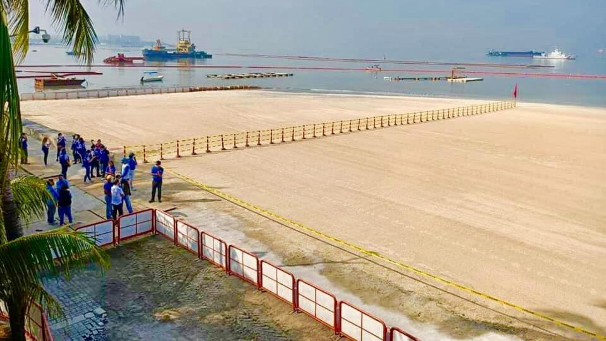Workers in blue uniforms gather near the fenced-off artificial Dolomite Beach at Manila Bay, Philippines, with calm waters and ships anchored offshore in the background.