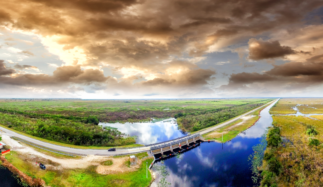 Aerial view of a road and waterway cutting through vast wetlands at Everglades National Park under dramatic skies.