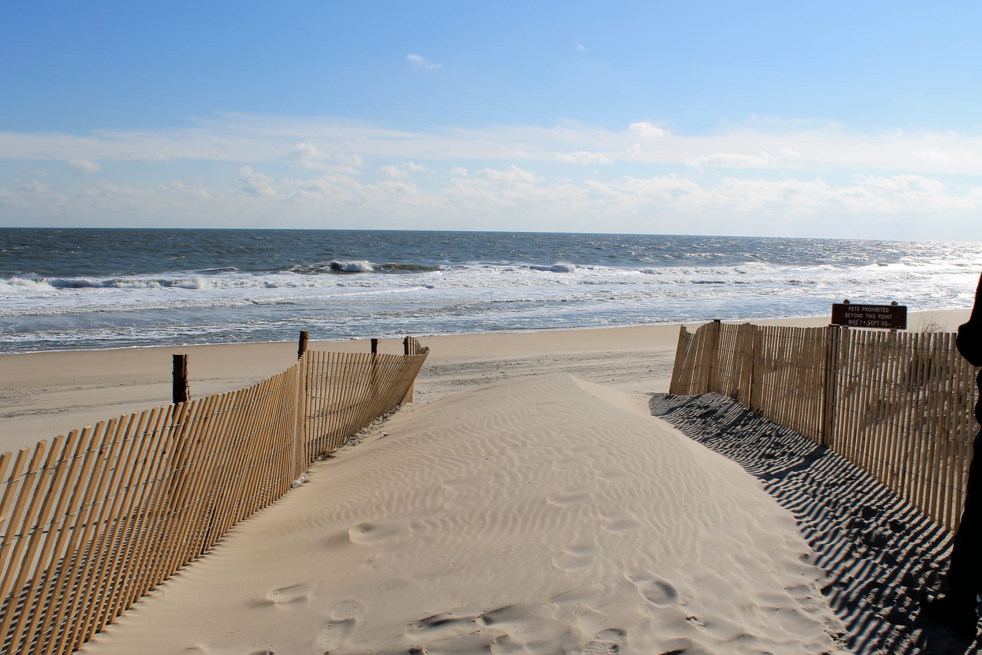 Wooden dune fencing along a sandy path leading to the ocean at Fenwick Island State Park, Delaware, with waves and a partly cloudy sky