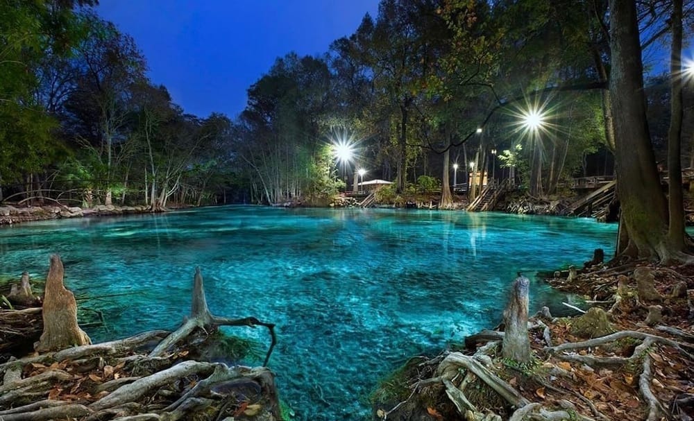 Nighttime view of Ginnie Springs in Florida with glowing turquoise water, tree roots in the foreground, and soft lights illuminating the surrounding cypress forest and wooden staircases.