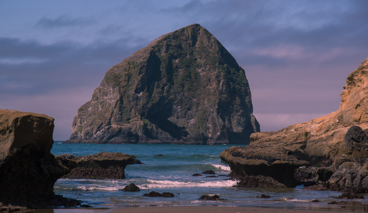Massive sea stack known as Haystack Rock rising from the ocean off Cape Kiwanda on the Oregon Coast, with rugged foreground rocks, calm waves, and a partly cloudy sky.