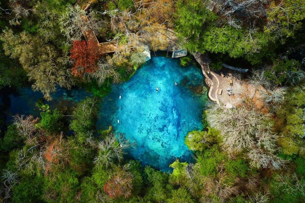 Aerial image of Ichetucknee River in Florida, featuring a vibrant blue spring pool encircled by dense forest and a wooden boardwalk leading to the water.