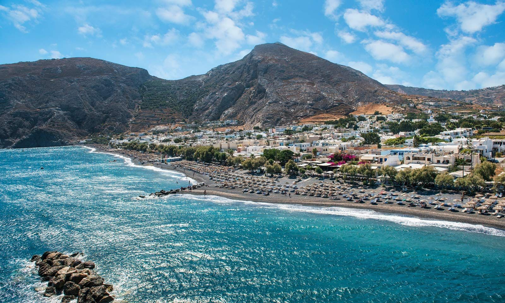 Coastal view of Kamari Beach in Santorini, Greece, showing black sand, rows of beach umbrellas, a vibrant seaside village, and dramatic mountain cliffs under a partly cloudy sky.