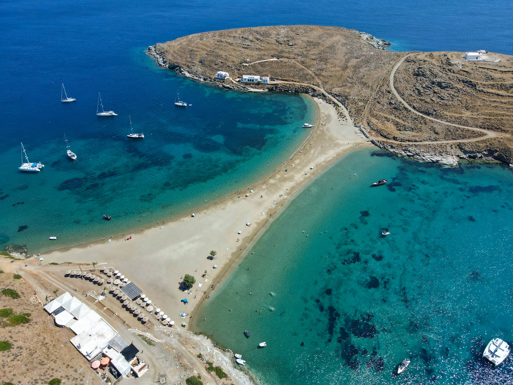 Aerial view of Kolona Beach on Kythnos Island, Greece, showing a narrow sandbar connecting two landmasses, with yachts anchored in clear blue waters on both sides.