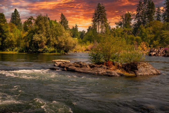 Scenic view of a calm river flowing through LaPine State Park in Oregon, with lush green trees and a dramatic orange and purple sunset sky in the background.