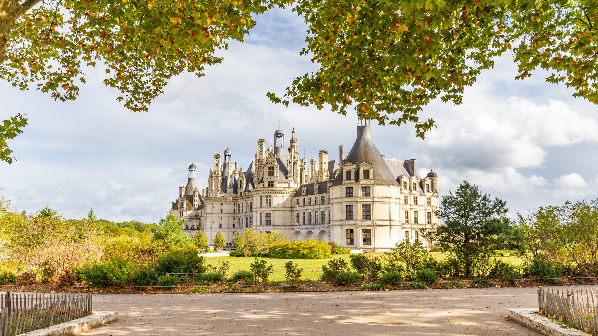 Château de Chambord in France’s Loire Valley, surrounded by fall foliage and green gardens under a partly cloudy sky.