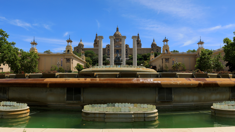 View of the Magic Fountain of Montjuïc in Barcelona with the National Art Museum of Catalonia in the background under a clear blue sky.
