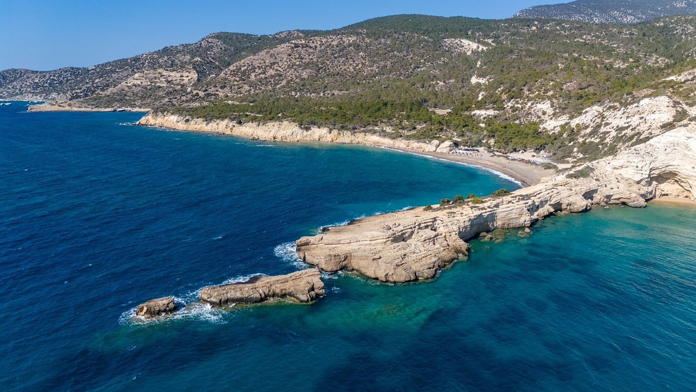 Aerial view of Monolithos Beach in Santorini, Greece, featuring a rocky peninsula stretching into the Aegean Sea, bordered by forested hills and a quiet sandy bay in the background.