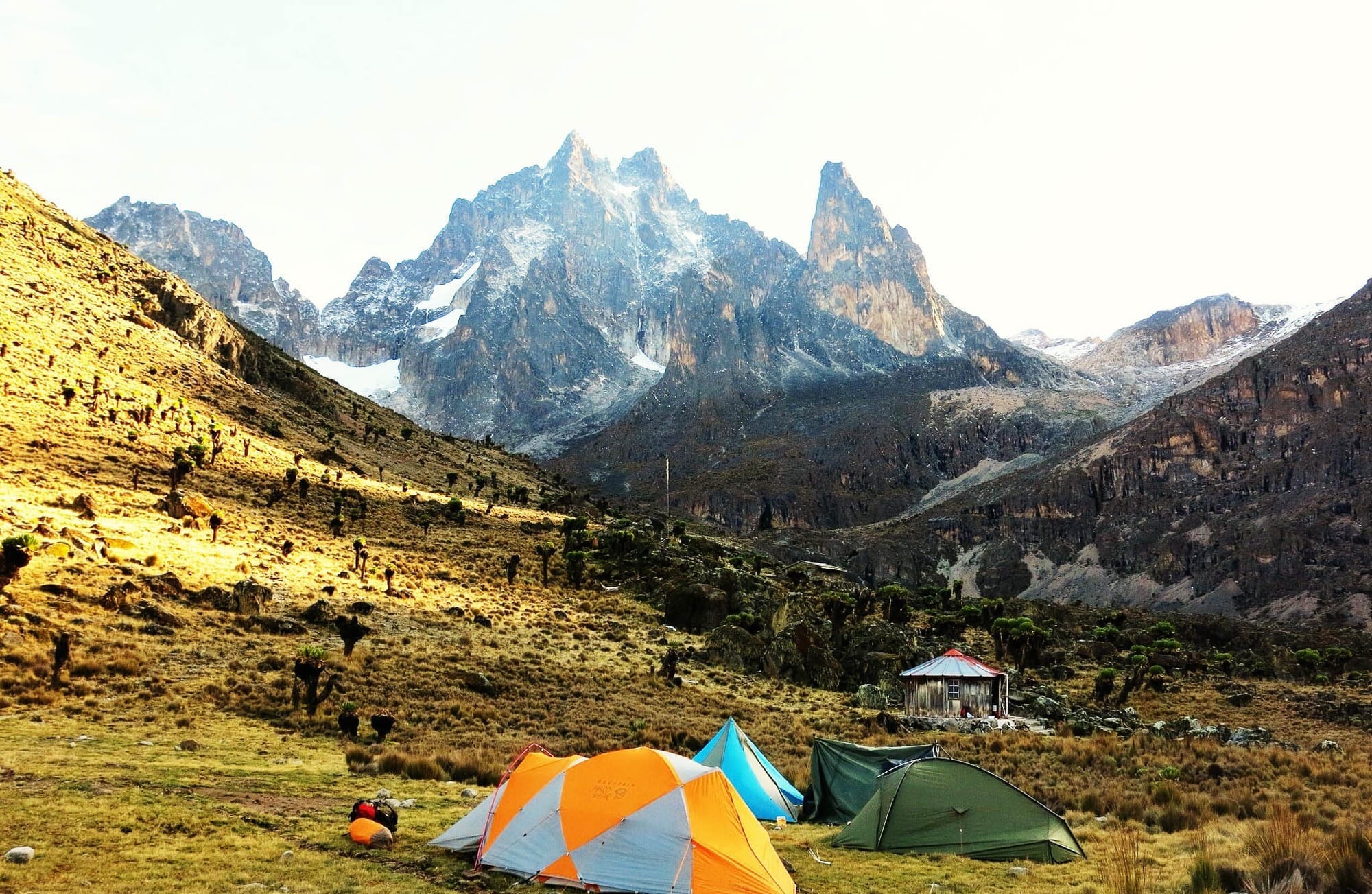 Colorful camping tents set up on a grassy valley beneath the rugged snow-capped peaks of Mount Kenya, with early morning sunlight casting shadows across the landscape.