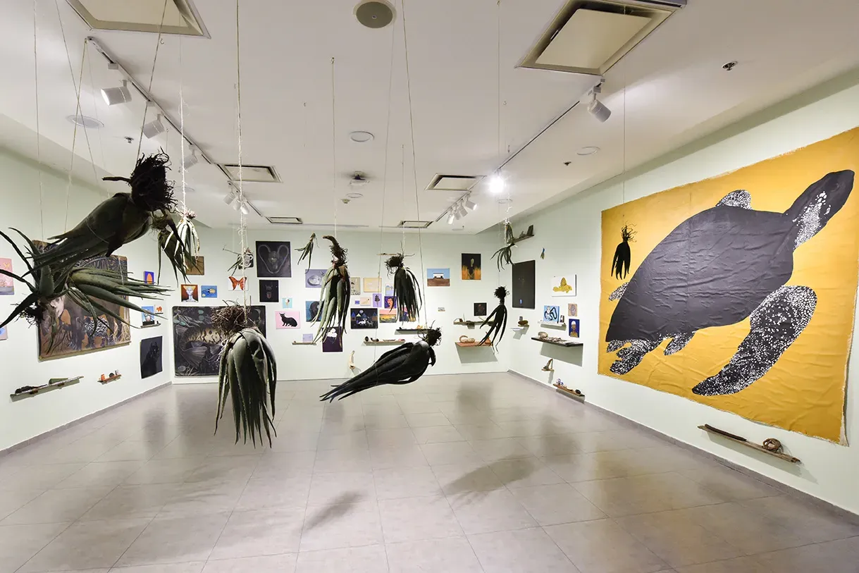 Contemporary art installation at the Museo de Arte Moderno de Bogotá, featuring suspended plant-like sculptures, eclectic wall art, and a large turtle mural on a yellow background.