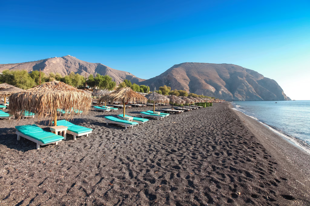 A wide-angle view of Perissa Beach in Santorini, Greece, showing black volcanic sand, rows of turquoise sunbeds under straw umbrellas, and a mountainous backdrop against a clear blue sky.
