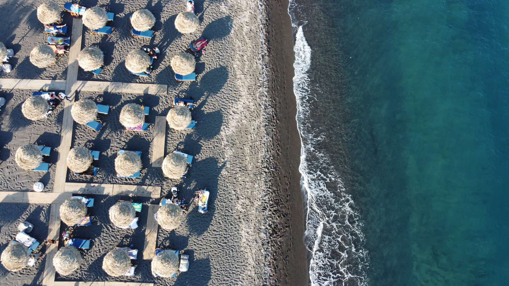 Aerial view of Perivolos Beach in Santorini, Greece, showcasing organized rows of sunbeds and umbrellas on dark sand next to gently rolling waves along the shoreline.