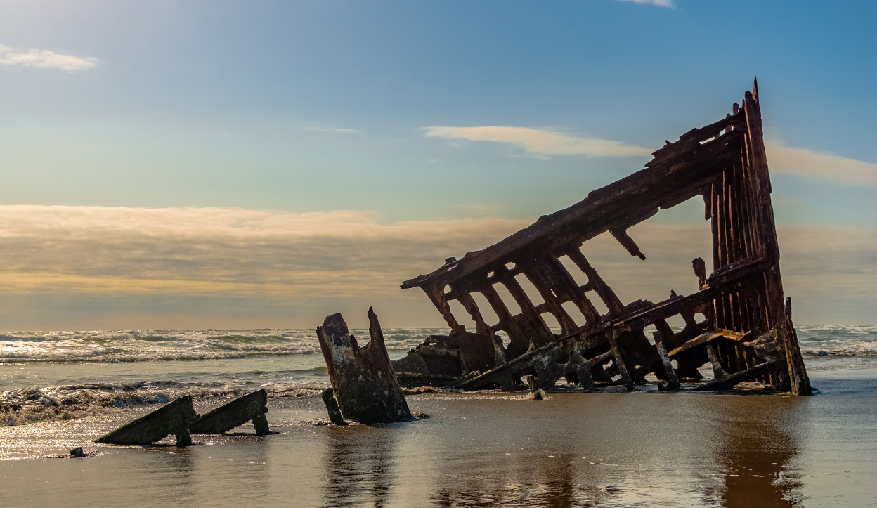 Rusted remains of the Peter Iredale shipwreck sitting on a sandy beach at Fort Stevens State Park in Astoria, Oregon, with ocean waves and a partly cloudy sky in the background.