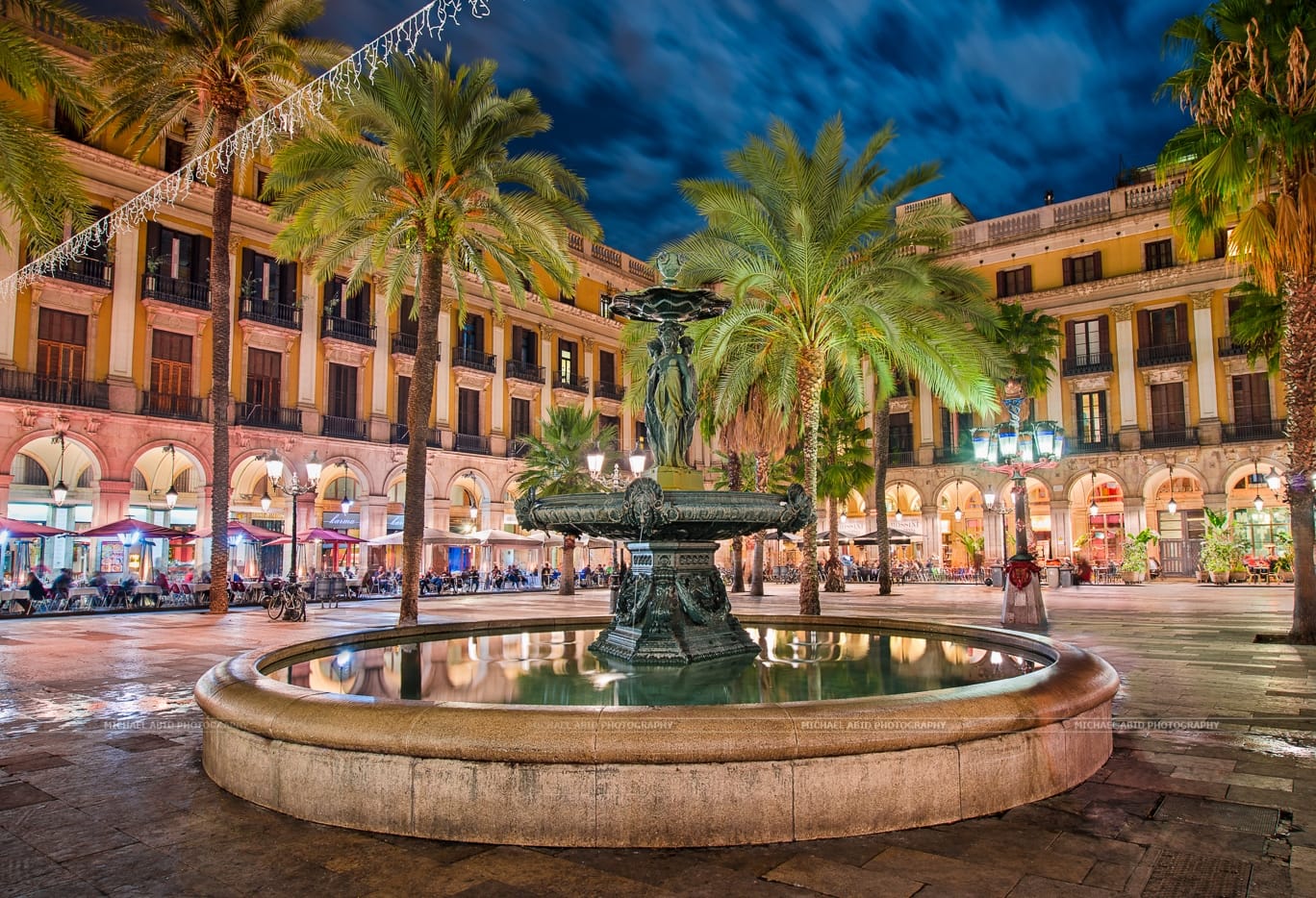 Plaça Reial at night with palm trees, glowing lights, and a central fountain surrounded by lively restaurants in the Gothic Quarter.