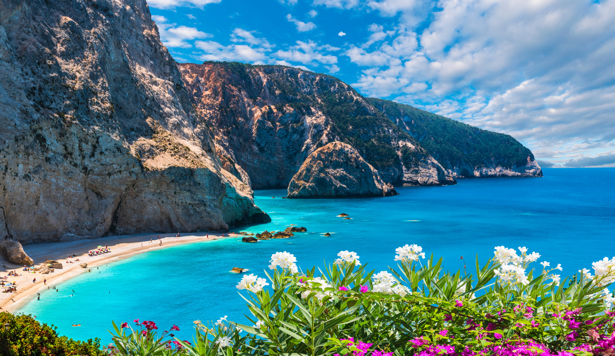Scenic view of Porto Katsiki Beach in Lefkada, Greece, with towering white cliffs, turquoise water, and blooming flowers in the foreground.