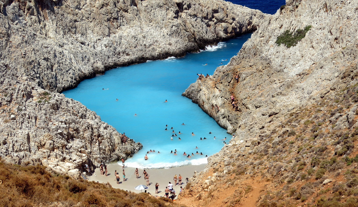 Seitan Limania Beach on Crete, Greece, featuring a narrow, rocky inlet filled with vivid blue water and surrounded by steep cliffs, with swimmers enjoying the natural cove.