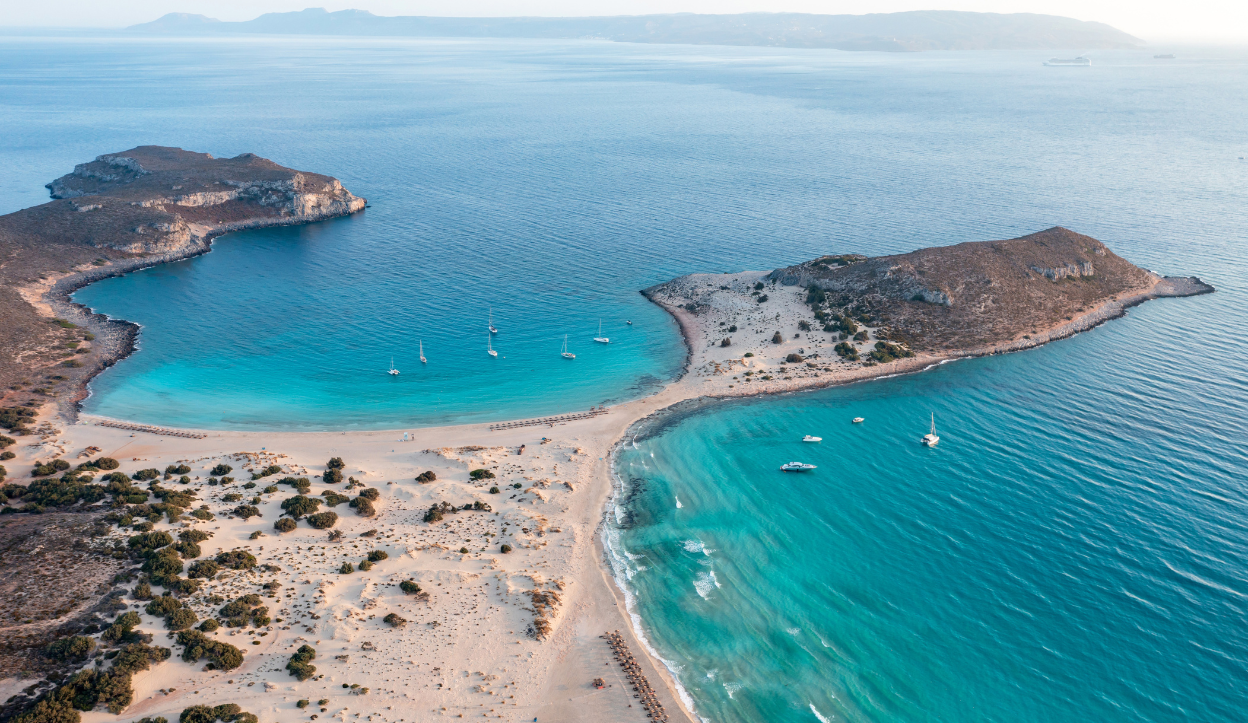 Aerial perspective of Simos Beach in Elafonisos, Greece, showcasing a double crescent shoreline with turquoise bays on either side of a sandy peninsula.