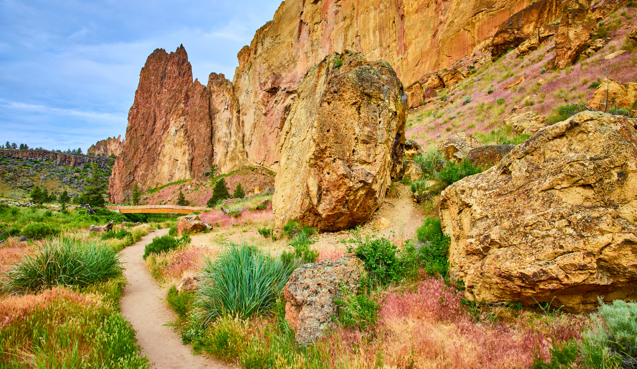 Dramatic desert landscape of Smith Rock State Park in Oregon featuring a winding dirt trail, towering red rock formations, and dry grasses and shrubs beneath a soft blue sky.