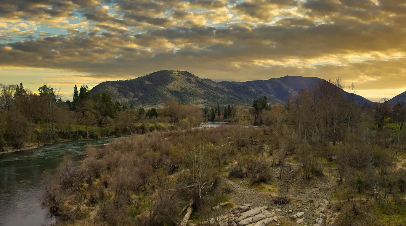Panoramic view of Valley of the Rogue State Park in Oregon, showcasing a winding river, leafless trees, and forested hills beneath a moody, cloud-filled sky at sunset.