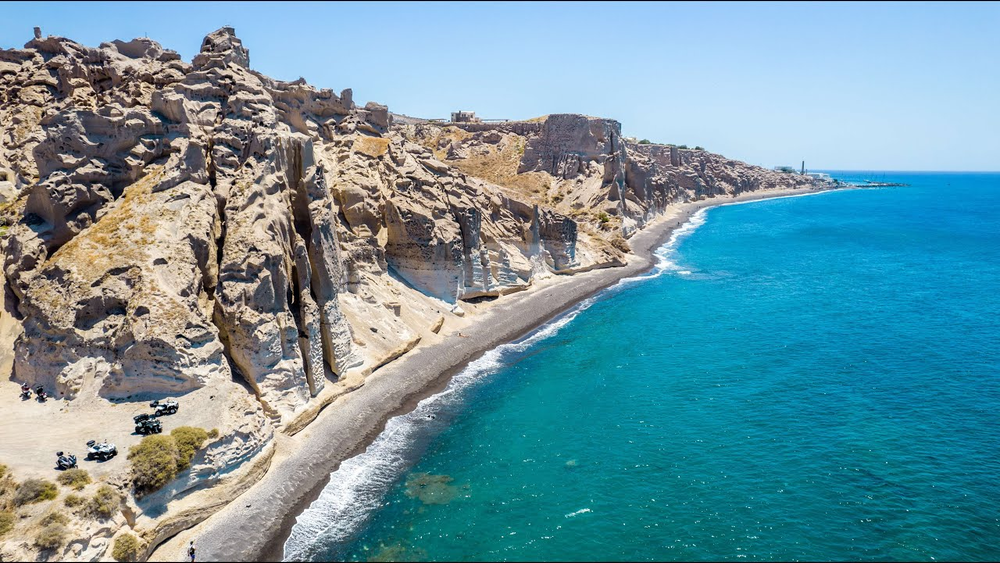 Aerial view of Vlychada Beach in Santorini, Greece, showcasing dramatic, sculpted white cliffs formed by volcanic ash, a narrow strip of dark sand beach.