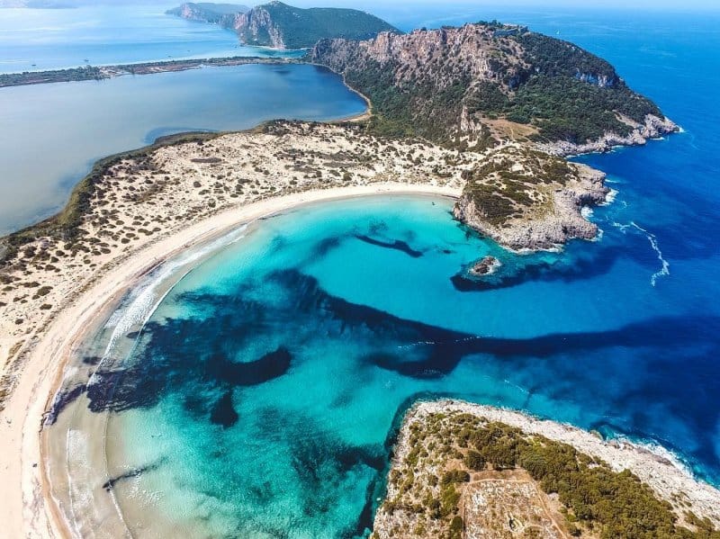 Drone view of Voidokilia Beach in the Peloponnese, Greece, with a perfectly curved sandy bay, turquoise waters, and a lagoon in the background.