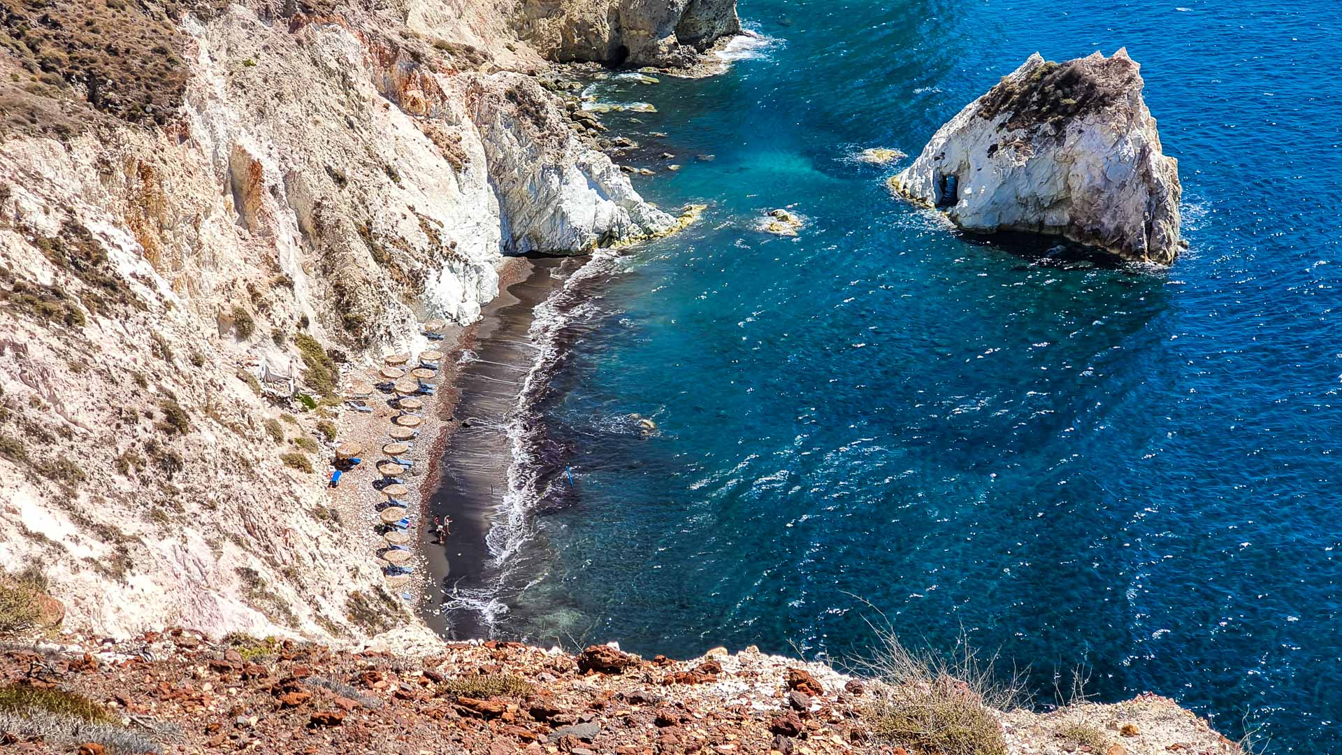 Aerial view of Santorini’s White Beach, nestled between dramatic white limestone cliffs and the deep blue Aegean Sea, with rows of beach umbrellas lining the narrow black sand shoreline.