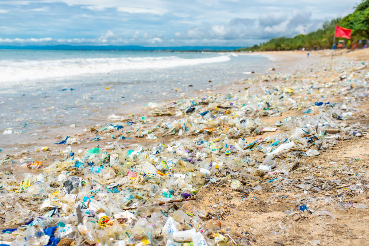 Plastic waste and debris littering the shoreline of a Kuta beach in Bali, Indonesia, with ocean waves washing more trash ashore and a red warning flag in the distance.