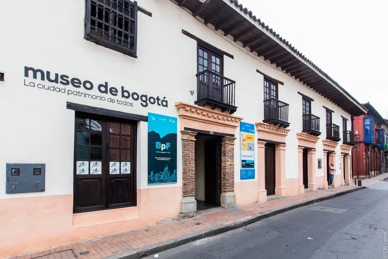 Exterior of the Museo de Bogotá in Colombia, housed in a colonial-style building with wooden balconies and cultural posters displayed along the sidewalk.