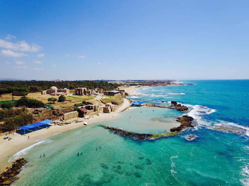 Aerial view of Achziv Beach near Nahariya, Israel, showing turquoise Mediterranean waters, rocky tidal pools, and sandy shores with historic stone structures in the background under a clear blue sky.