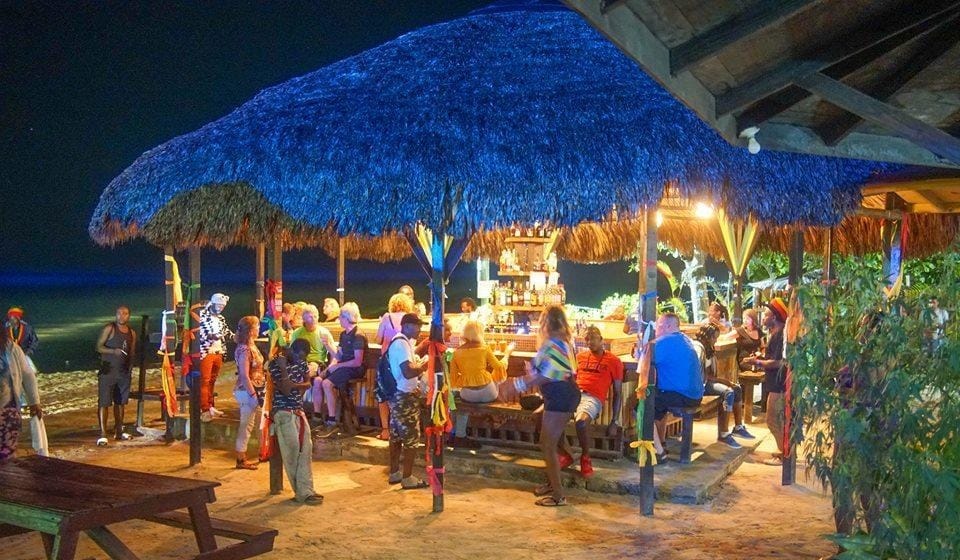 Beachside thatched roof bar at night with people enjoying drinks and socializing at Bourbon Beach in Negril, Jamaica. 