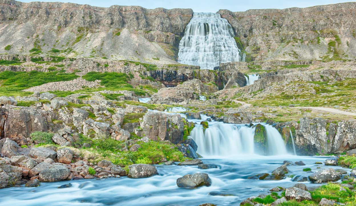 Wide-angle view of Dynjandi waterfall cascading down terraced rock formations in the Westfjords of Iceland, surrounded by mossy cliffs, rocky terrain, and clear blue streams in the foreground.
