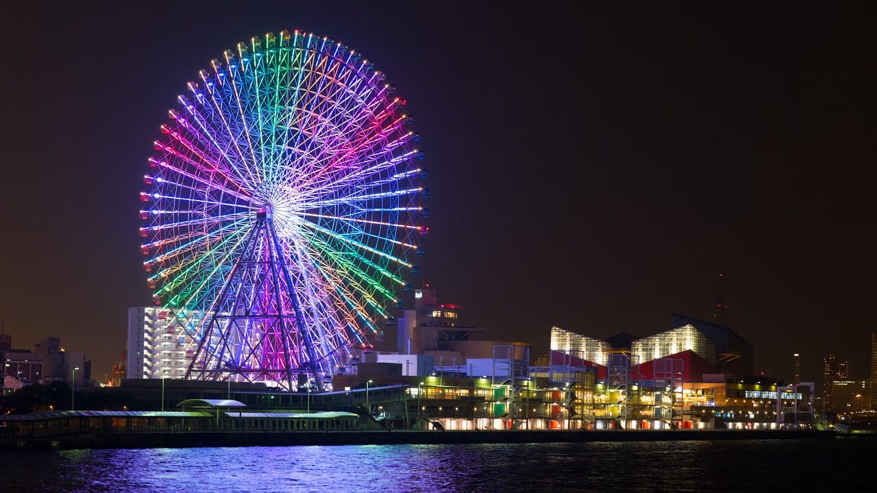 A giant Ferris wheel on the water front in Osaka Japan all lit up in bright colors at night.