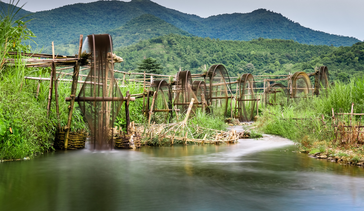Traditional bamboo water wheels line a small river in a lush rural area of Vietnam, with forested mountains in the background and vibrant green vegetation surrounding the flowing stream.