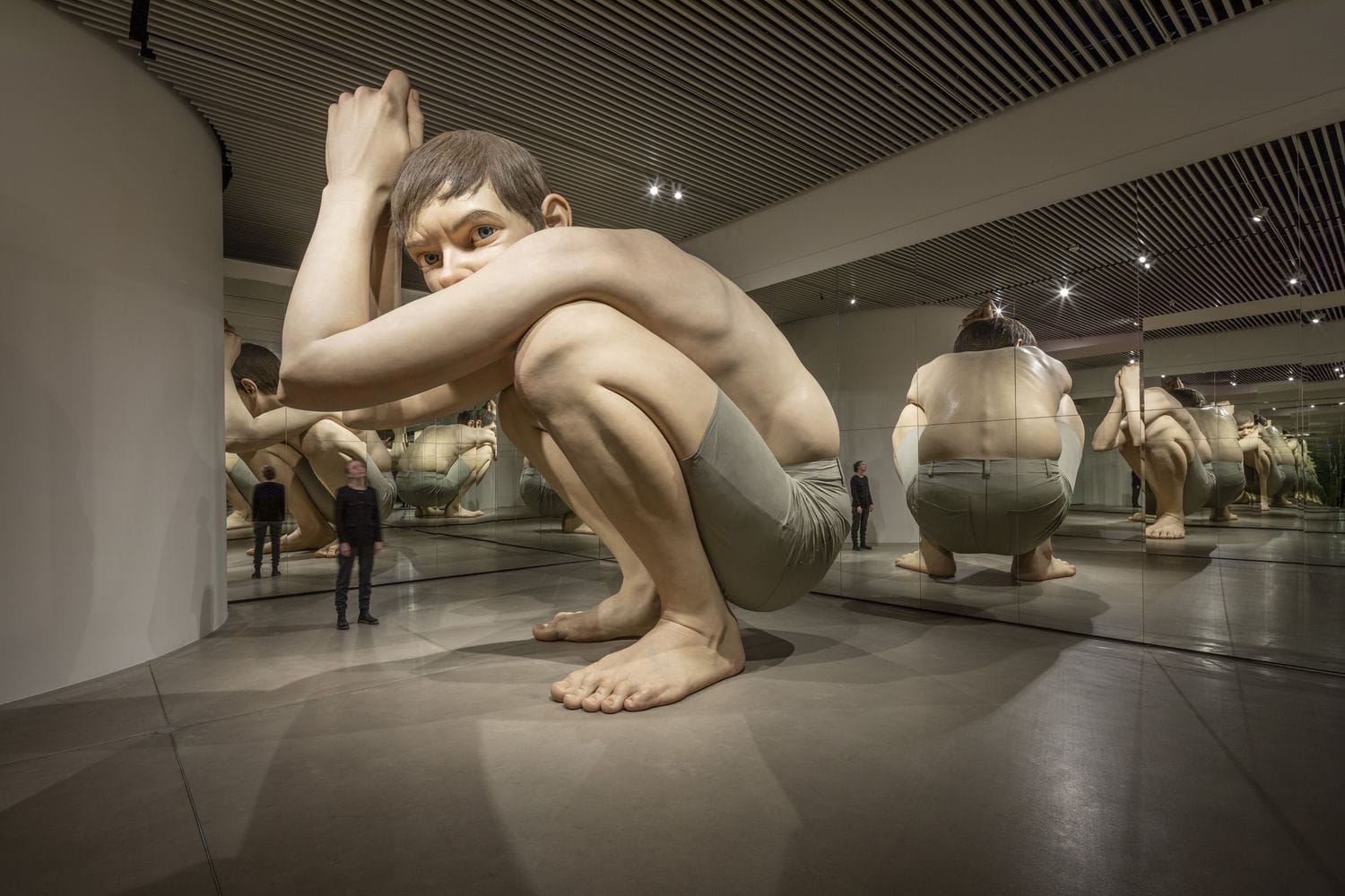 A giant hyperrealistic sculpture of a crouching boy inside ARoS Aarhus Art Museum in Denmark, with museum visitors standing nearby for scale.