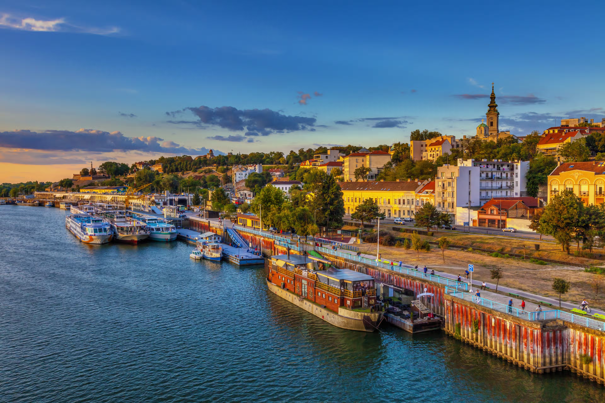 A river running through Serbia with long boats anchored next to the shore.