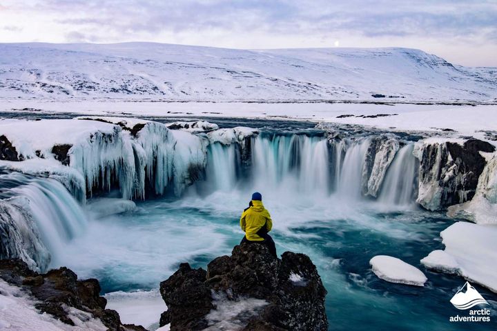 Man sitting on a rock looking at a horseshoe shaped waterfall in Iceland with snow and ice covering the landscape