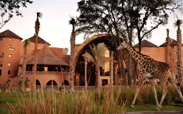 A Giraffe walking on the grass in front of a tan colored resort building with a glass front entrance. 