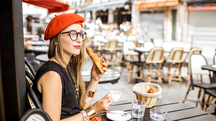 A woman sitting at a cafe in Paris, wearing a red Parisian hat and eating a bagel. 